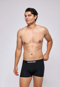 Shirtless man adjusts black boxer briefs with “SNOCKS” waistband, standing against plain light gray background.