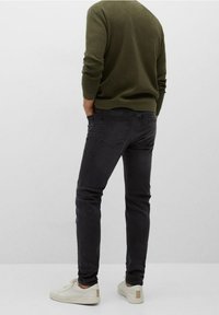 Mu muž v tmavě šedých slim-fit džínech, olivově zeleném svetru a bílých teniskách, stojící s rukama v kapsách na jednobarevném pozadí.