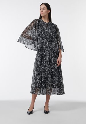 Vrouw draagt een zwarte midi-jurk met doorschijnende, brede vliegende mouwen en witte abstracte vlekken, gecombineerd met zwarte puntige pumps, staand.