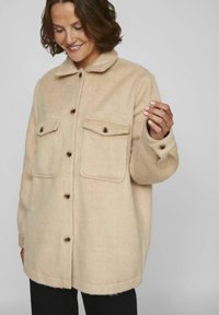 Beige villasekoituksessa oleva takki-paita, jossa on terävä kaulus, etupuolella nappikiinnitys, kaksi rintataskua ja rento istuvuus. Varustettu puisilla napilla.