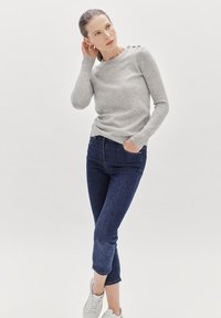 Suéter de punto gris claro con cuello redondo y acentos de botones en el hombro, combinado con jeans recortados azul oscuro y zapatillas blancas.