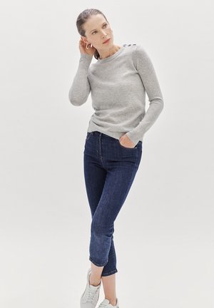 Lichtgrijze gebreide sweater met ronde halslijn en knoopaccenten op de schouder, gecombineerd met donkerblauwe cropped jeans en witte sneakers.