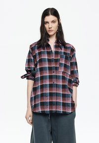 Camisa de cuadros en rojo, negro y azul con botones frontales, bolsillos y mangas remangadas, hecha de una tela suave y texturizada.
