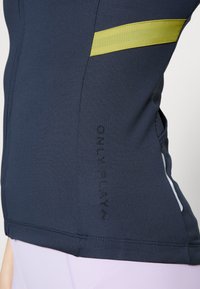 Haut de sport marine avec une bande jaune, tissu texturé et impression noire "ONLY PLAY" sur le côté. Présente un bord contrastant blanc à l'ourlet.