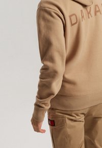 Persona vistiendo una sudadera marrón claro con el texto "DAKAR" y pantalones a juego del mismo color con un parche en el bolsillo lateral, posando frente a un fondo liso.