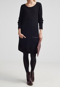Robe en maille noire à manches longues, coupe décontractée et deux poches avant. Portée avec des collants noirs et des bottines marron, tenant une pochette bordeaux.