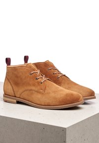 Botas chukka de ante marrón con cordones marrones, punta redonda y suela de perfil bajo. Presenta una pequeña pestaña en el talón y un sutil logo de marca.