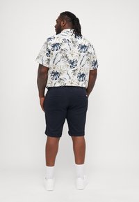 Camisa de manga corta con un patrón de hojas tropicales en verde y azul, combinada con pantalones cortos oscuros de color marino, y llevada con zapatillas blancas y calcetines.