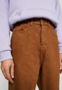 Brązowe bawełniane spodnie o teksturowanym wykończeniu, z przodem zapinanym na guziki i bocznymi kieszeniami. Zestawione z jasnofioletową bluzą.