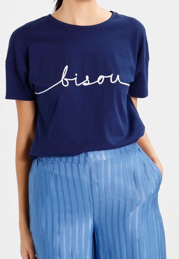 Žena oblečená v námořnickém modrém tričku s textem "bisou" a světle modrých širokých pruhovaných kalhotách, s rukama v kapsách, na bílém pozadí.