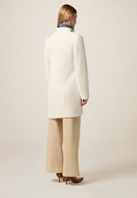 Abrigo de lana blanca texturizada con un cuello alto, detalles de botones y un corte holgado, combinado con pantalones de pierna ancha beige y tacones marrones.