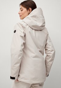 Beige vandtæt jakke med hætte, der har en sort knapdetalje på ærmet og et hvidt logo på bagsiden. Glat tekstur.