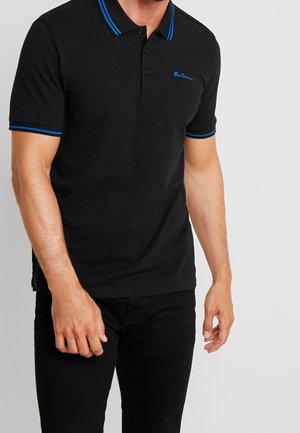 Mann trägt ein schwarzes Polohemd mit blauer Einfassung am Kragen und an den Ärmeln, kombiniert mit schwarzer Hose, stehend vor einem schlichten hellen Hintergrund.