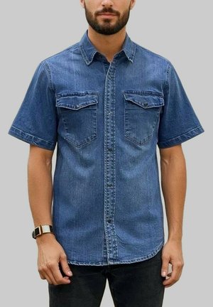 Homme portant une chemise en denim bleue à manches courtes avec deux poches poitrine et un pantalon noir, sur un fond gris uni.