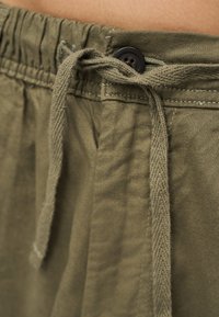 Nærbillede af olivengrønne bukser med snøre, elastisk linning og sort knap foran.