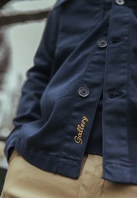 Tumši zila kokvilnas tvila krekla ar pogām, ar zelta izšuvumu "Gallery" detaļu un pelēkām pogām. Novietota uz kakijas krāsas biksēm.