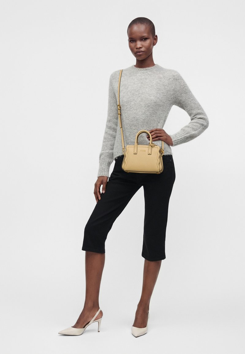 Femme portant un pull gris, un pantalon noir aux genoux, des talons beiges et portant un petit sac à main beige avec une bandoulière.