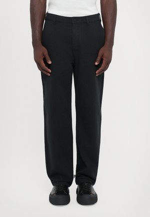Schwarze gerade geschnittene Hosen aus einem glatten Stoff mit einem Knopfverschluss vorne. Getragen mit schwarzen Turnschuhen. Einfaches Design, keine sichtbaren Muster.