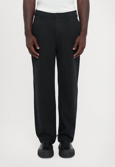 Pantalon noir à jambe droite en tissu lisse avec une fermeture à bouton à l'avant. Porté avec des baskets noires. Design simple, sans motifs visibles.