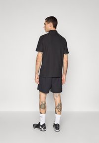 Sort polo-shirt med korte ærmer, parret med mørke shorts. Individet bærer sorte sneakers og hvide sokker, og viser tatoveringer på benene.