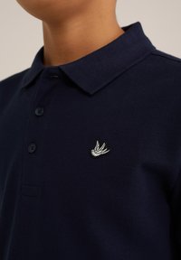 Laivyno spalvos polo marškiniai, pagaminti iš tekstūruoto audinio, turintys klasikinį apykaklę, tris mygtukus ir baltą siuvinėtą paukščio logotipą kairėje krūtinės pusėje.