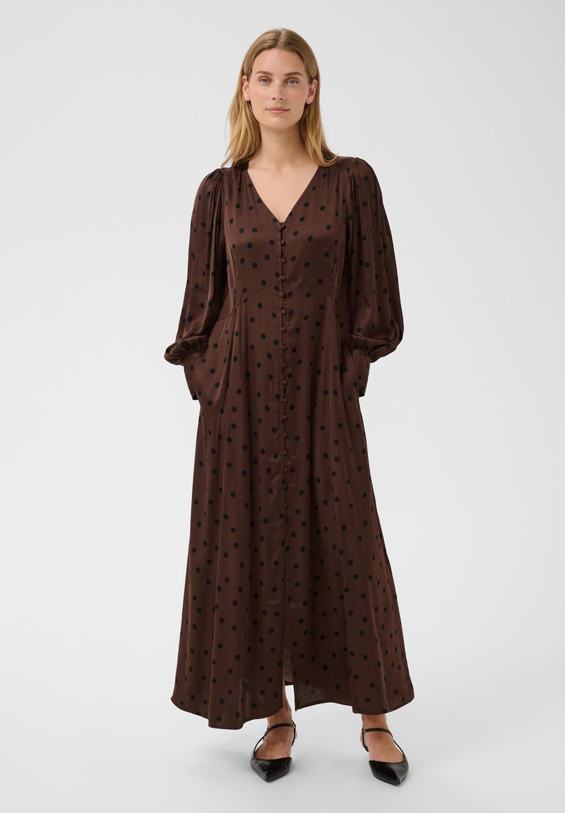 Femme portant une longue robe marron à pois noirs, manches longues bouffantes, boutons à l'avant et ballerines noires pointues, debout sur un fond blanc.