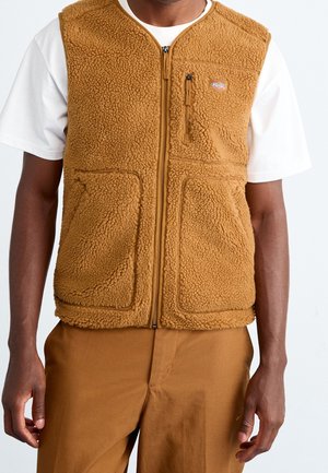 Homme portant un gilet zippé marron duveteux sur une chemise blanche à manches courtes et un pantalon marron assorti, debout devant un fond uni.