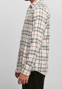 Man in een wit shirt met lange mouwen en een zwart-rood geruit patroon, staand in profil tegen een effen achtergrond.