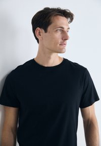 Young man with short brown hair and blue eyes wearing a black t-shirt, looking to the right against a plain white background.