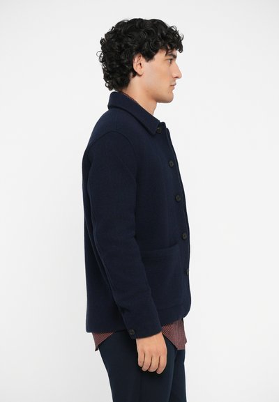 Veste en laine bleu marine avec un devant boutonné, un col pointu, deux poches latérales et une finition texturée, portée sur une chemise à motif.
