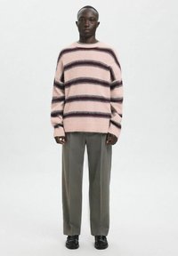 Dikke roze trui met donker kastanjebruine en grijze strepen, ronde hals en geribbelde manchetten, gecombineerd met lichtgrijze, wijde pijpen broek en zwarte schoenen.