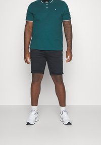 Ciemnozielona koszulka polo z białymi wykończeniami, czarne szorty dżinsowe oraz białe sneakersy, z podwiniętymi nogawkami i widocznym logo marki na koszulce.