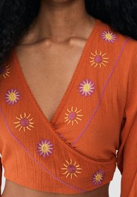 Rostfärgad omslagsblus i texturerat tyg, med broderade solmotiv i gult och lila. V-ringad design med accentstygning.