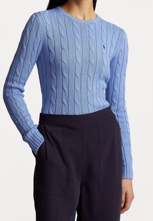 Femme portant un pull en maille torsadée bleu clair et un pantalon taille haute foncé, une main dans la poche, sur un fond uni.