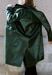 Cappotto in pelle verde scuro con design strutturato, dotato di spacco posteriore, texture liscia e tasche laterali. Indossato aperto per mostrare l'interno.
