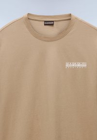 Béžové tričko s kulatým výstřihem s bílým logem "Napapijri geographic" na levé straně hrudi, viditelné prošití na ramenech a kolem výstřihu.