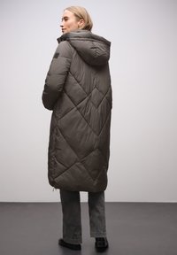 Manteau rembourré vert olive avec motif matelassé en forme de losanges, capuche et fermeture éclair à l'avant. Porté avec un pantalon gris et des chaussures noires.