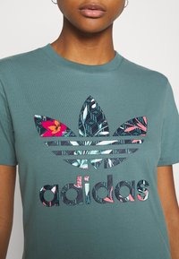 Grön bomullströja med en stor, flerfärgad Adidas-logotyp med geometriska och florala mönster. Kortsvarm  ärmar och rundad hals.