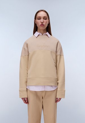 Jonge vrouw met stijlbruin haar, gekleed in een gelaagde beige trui over een roze overhemd met kraag en beige broek, staand tegen een lichte achtergrond.