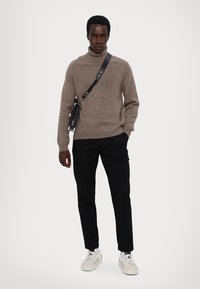 Bruine wollen turtleneck trui, zwarte broek en witte sneakers. Zwarte schoudertas met zilveren accenten, staande tegen een effen achtergrond.