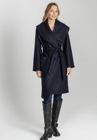 Niebieski płaszcz wełniany z szerokim kołnierzem, pasującym paskiem i dwiema przednimi kieszeniami. Modelka połączyła go z czarnymi botkami do kolan i dżinsami.