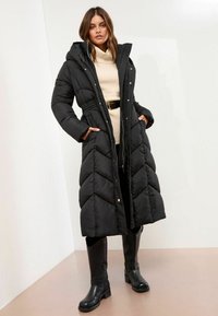 Cappotto lungo nero imbottito con trapuntatura a zig zag, cappuccio e vita regolabile con cordoncino. Abbinato a stivali neri alti fino al ginocchio e a un maglione beige.