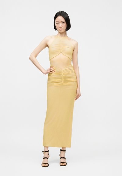 Γυναίκα που φοράει ανοιχτό κίτρινο, μανίκless φόρεμα halter με ρυτίδες και κοψίματα στη μέση, στέκεται με το ένα χέρι στη μέση και μαύρα ψηλοτάκουνα σανδάλια με λουράκια.