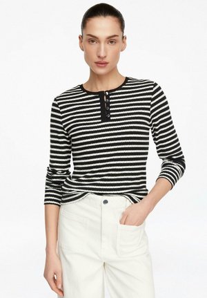 Kobieta z włosami związanymi do tyłu, ubrana w czarno-białą koszulę w paski z długim rękawem i guzikami oraz białe spodnie, z ręką w kieszeni.