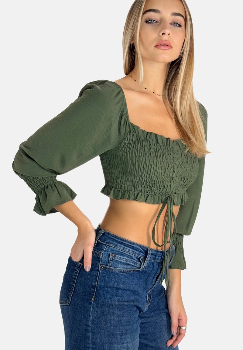 Olivgrünes Cropped-Top mit langen Puffärmeln, geraffter Textur, quadratischem Ausschnitt und verstellbarem Bindeband am Saum. Kombiniert mit blauen Jeans.
