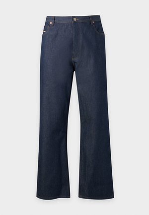 Junger Mann, der ein ärmelloses, geripptes hellgrünes Oberteil, dunkle Jeans, schwarze Loafers und einen über die Schultern gelegten grünen Pullover trägt.