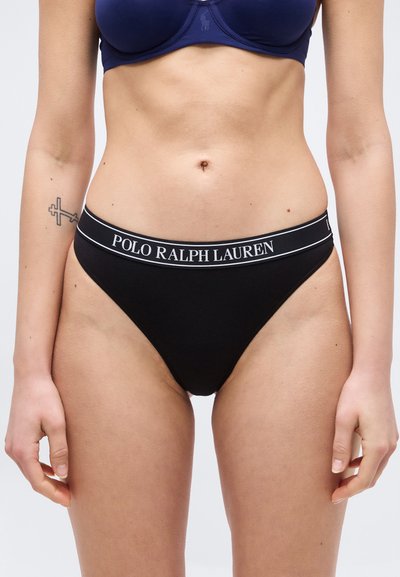 Γυναίκα που φοράει μαύρα εσώρουχα Polo Ralph Lauren και μπλε σκούρο σουτιέν, με ορατό τατουάζ σταυρού στο αριστερό αντιβράχιο, στέκεται μπροστά σε απλό φόντο.