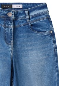 Gros plan sur la taille d'un jean en denim bleu avec bouton frontal, passants pour ceinture, poche et étiquette "Cecil Denim" à l'intérieur de la ceinture.