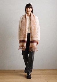 Wool scarf in soft pink with white floral patterns and dark stripes, fringed edges. Model wears an ivory textured jacket and dark pants.