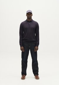 Sötétlila hosszú ujjú polo, közepes kötési struktúrával, sötét farmernadrággal és barna fűzős cipővel párosítva. Egy világoskék sapka egészíti ki a megjelenést.